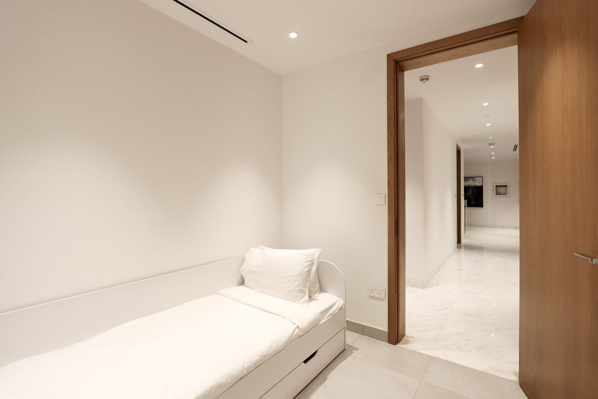 A minimalist bedroom with a single bed, a wooden door, and a hallway leading to another room.