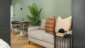Un salon avec un canapé gris, deux fauteuils, une petite table d’appoint sur laquelle se trouvent un livre et une télécommande, et une grande plante verte.