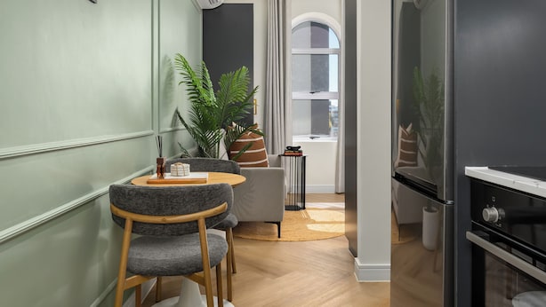 Une cuisine moderne dotée d’un four encastré, un coin repas avec une table ronde et des chaises grises, et une grande fenêtre avec des rideaux.
