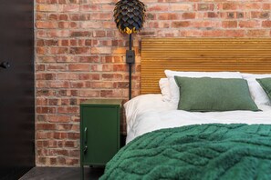 Een kamer met een bakstenen muur, een houten hoofdbord, een groen kastje en een bed met groene kussens.