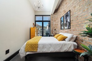 Een slaapkamer met een bed, een raam, een bakstenen muur en een kroonluchter.