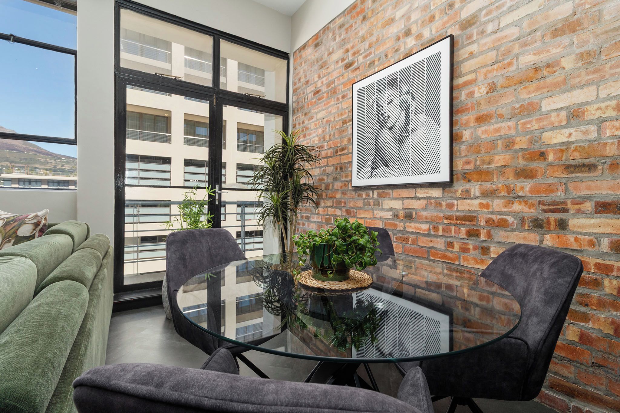 Un espacio habitable moderno con una mesa de vidrio, dos sillas, un sofá verde y una pared de ladrillo.
