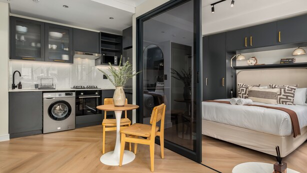 A modern kitchen with a washing machine, a dining area with a table and chairs, and a bedroom with a bed and bedside lamps. - The Valour Charming Apartment | City and Hill Views (Cape Town)