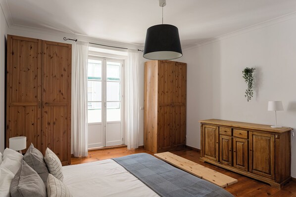 A bedroom with a bed, wooden wardrobe, a bedside table, a hanging plant, and a window with curtains.