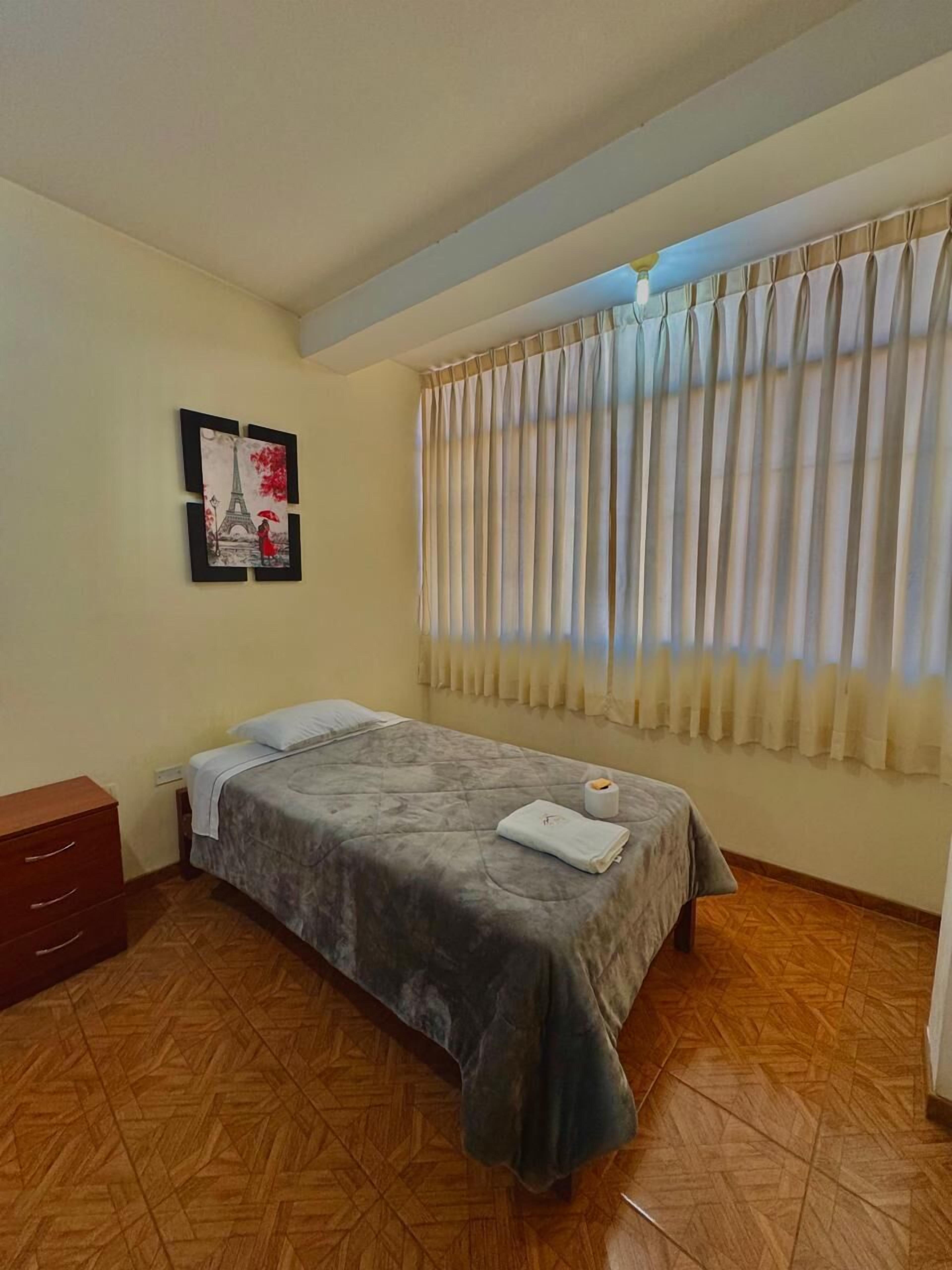 A single bed with a grey blanket, a wooden nightstand, a framed picture of the Eiffel Tower, and a window with beige curtains.