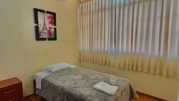 A single bed with a grey blanket, a wooden nightstand, a framed picture of the Eiffel Tower, and a window with beige curtains.