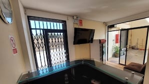 A reception area with a glass counter, a mounted television, and a view of the outside through a large window with a metal gate. - HOTEL CERRO BLANCO (Nazca)