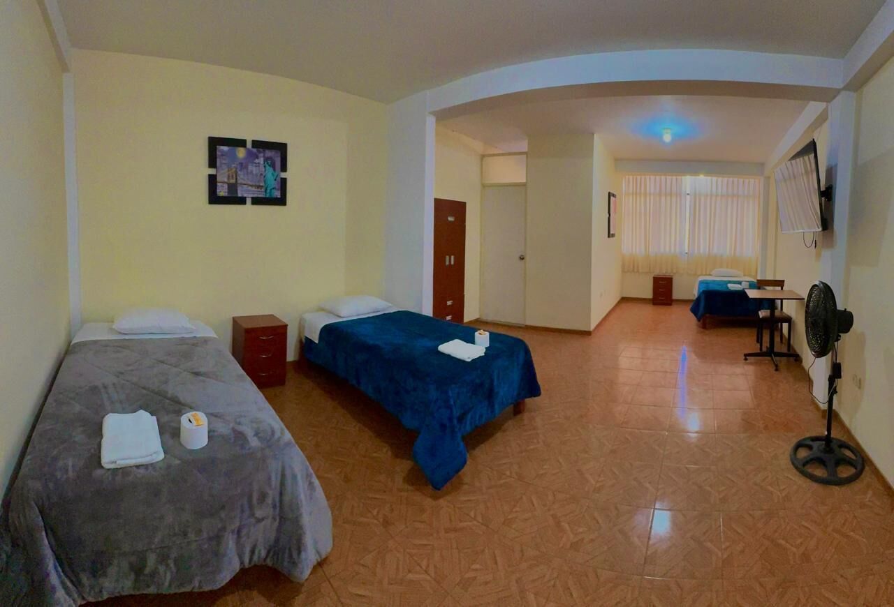 A hotel room with two single beds, a desk with a chair, a standing fan, and a TV mounted on the wall.