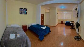 A hotel room with two single beds, a desk with a chair, a standing fan, and a TV mounted on the wall.