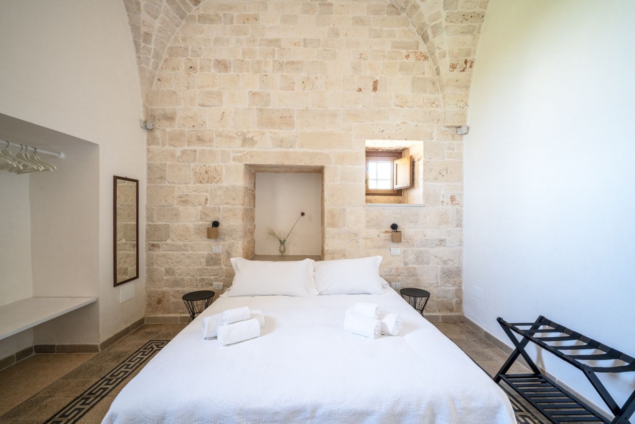 A bedroom with a stone wall, a bed with white linens, a small window, and a bench.