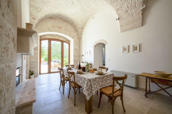 Un coin repas avec une table dressée pour quatre personnes, entouré de chaises en bois, d’une cheminée en pierre et offrant une vue à travers une porte en arc de cercle.