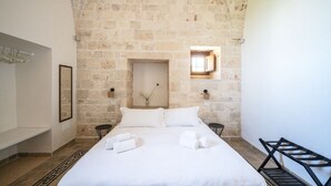 Ein Zimmer mit Steinwand, einem Bett mit weißer Bettwäsche, einem kleinen Fenster und einer Bank.