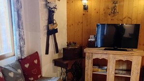 A cozy room with a wooden bench, a small table, a television on a wooden stand, and a wall-mounted shelf with decorative items. - Apartment in resort center chalet (Chamrousse)
