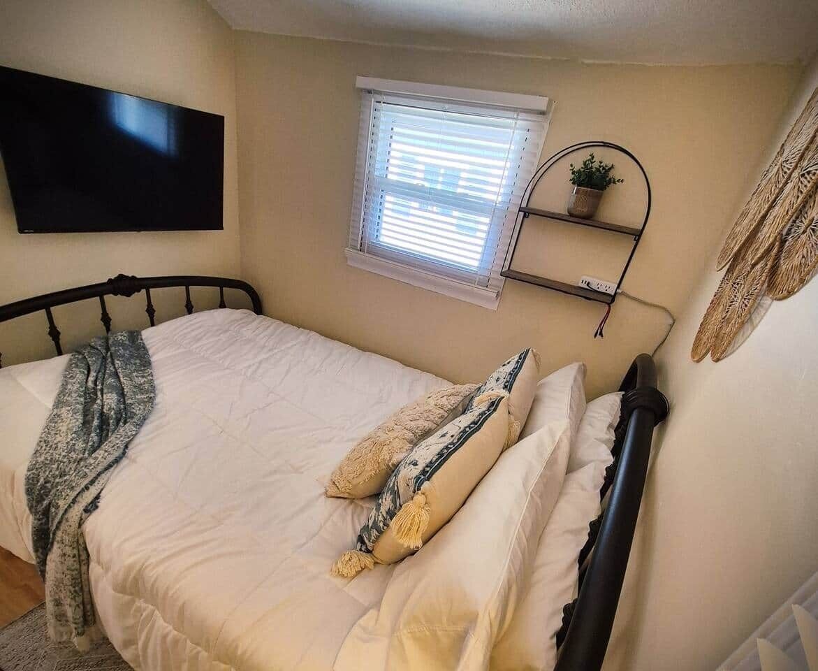A bedroom with a bed, a TV mounted on the wall, a window with blinds, and a shelf with a plant.
