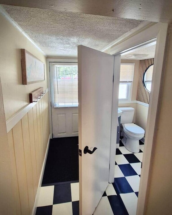 A narrow hallway with a black and white checkered floor, a white door, and a small window with blinds. - Parkview Boho Cottage 3 - Maple Meadows (Leamington)