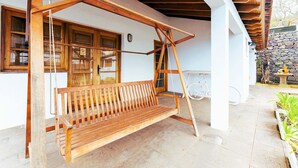 Un banc-balançoire en bois placé sous une structure en poutres de bois, un mur blanc avec une fenêtre et un mur en pierre avec une lanterne.
