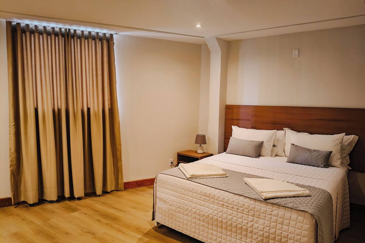 Una habitación de hotel con una cama grande, una mesita de noche, una lámpara y una ventana con cortinas.