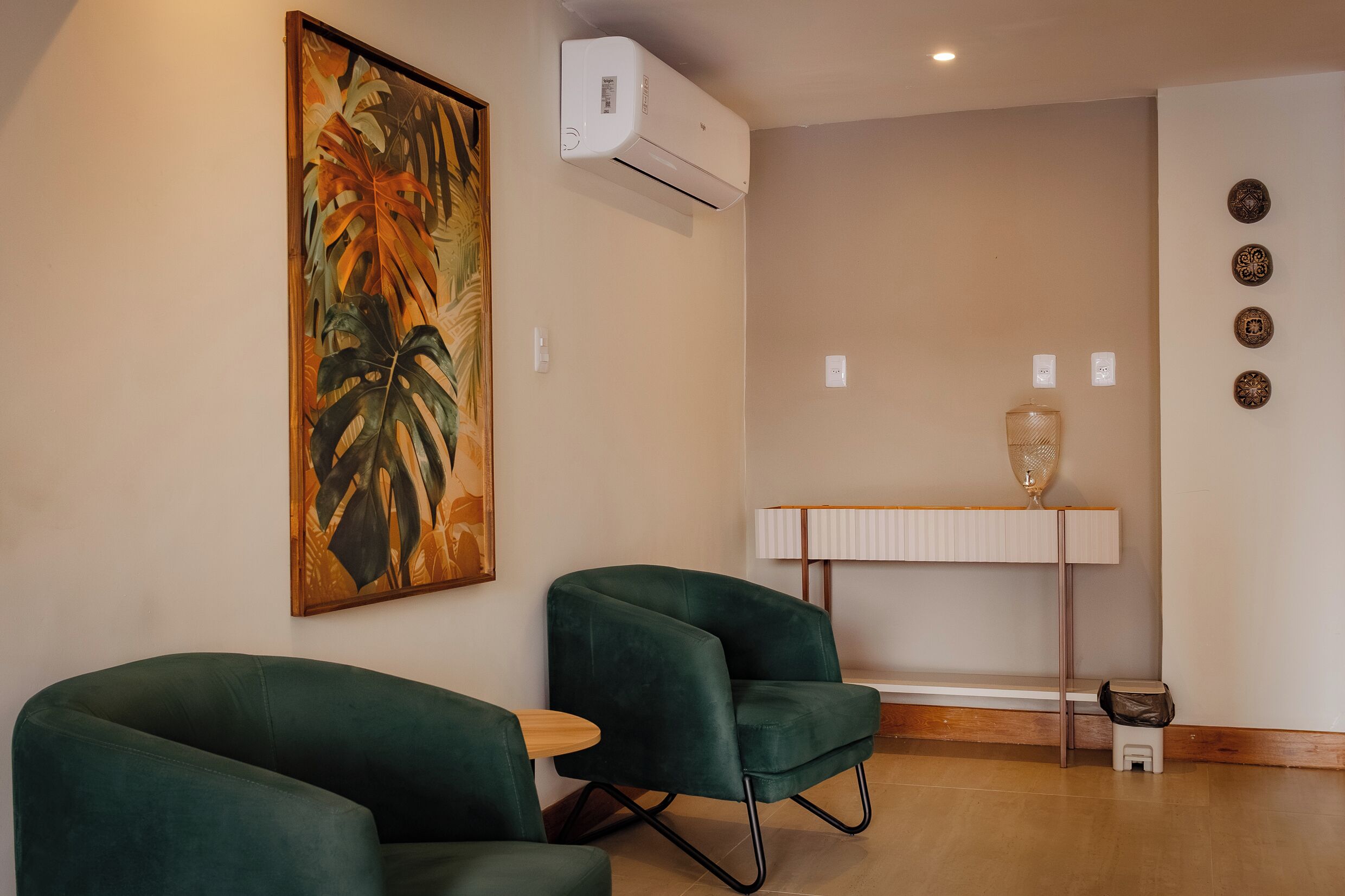 Una habitación con dos sillas verdes, una mesa, un cuadro, un aire acondicionado, una lámpara, un taburete pequeño y un adorno de pared.
