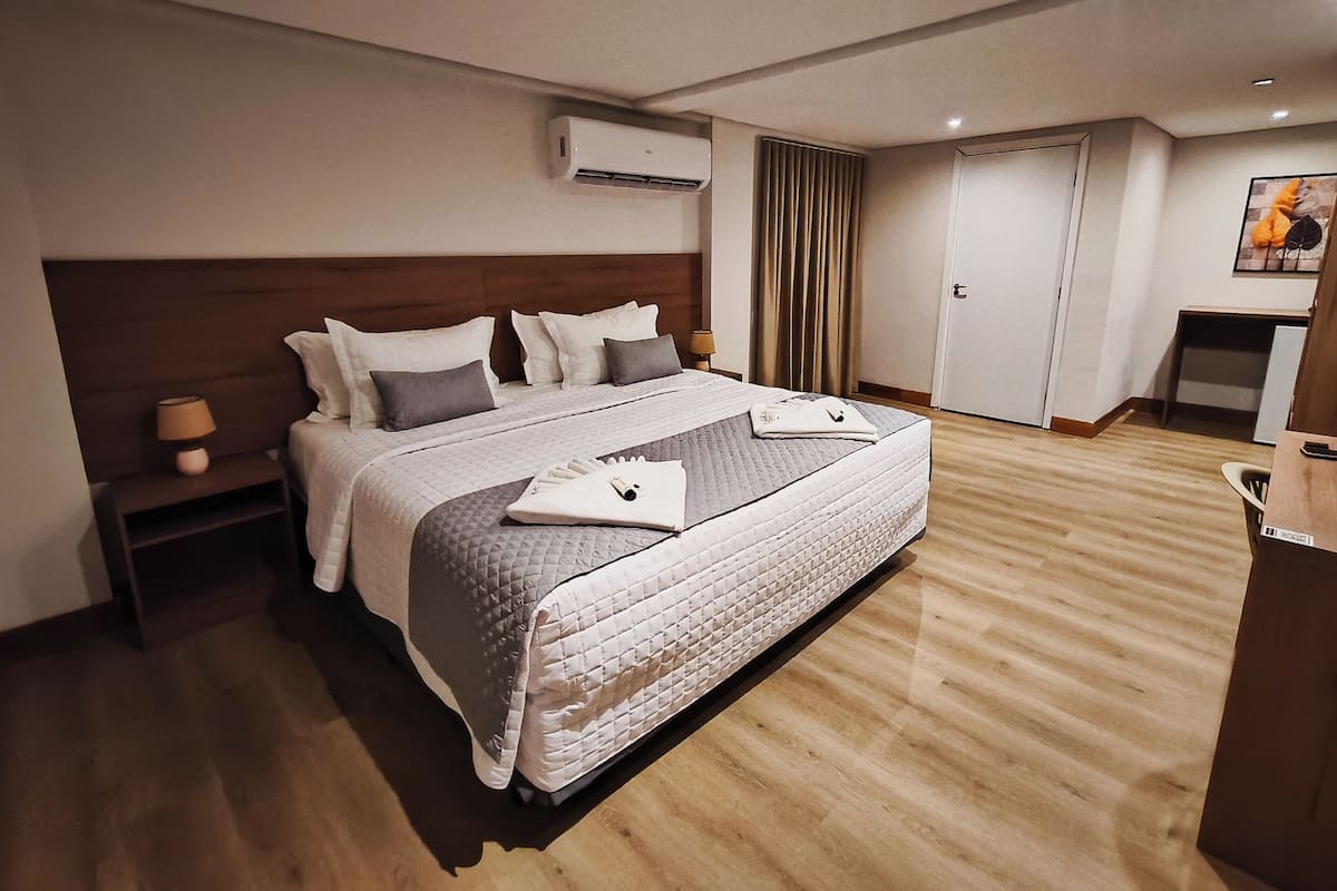 Habitación de hotel con una cama grande, una mesita de noche, un equipo de aire acondicionado, una puerta y un pequeño escritorio con una silla.