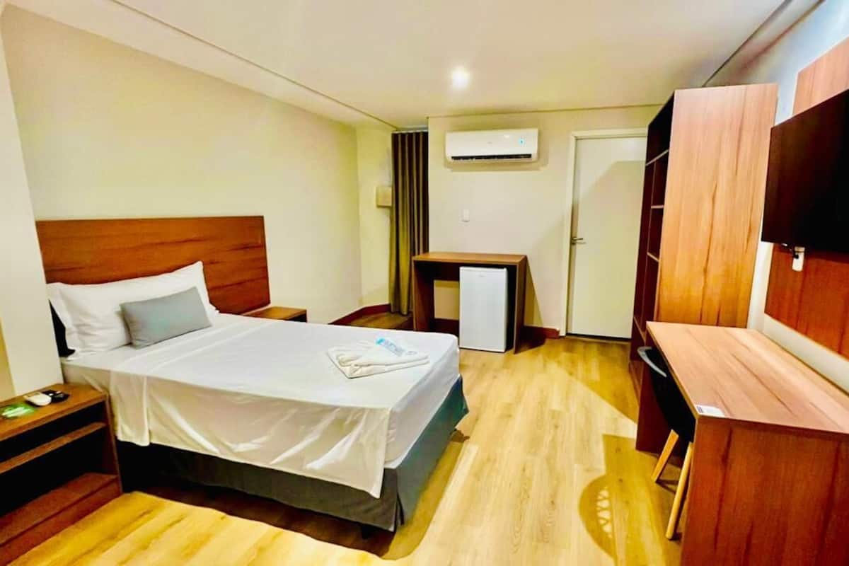 Habitación de hotel con cama, mesita de noche, escritorio, silla, televisor, armario y aire acondicionado.