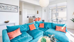 Un salon moderne avec un canapé d’angle turquoise, une table basse en bois et une vue sur la cuisine et l’espace repas.
