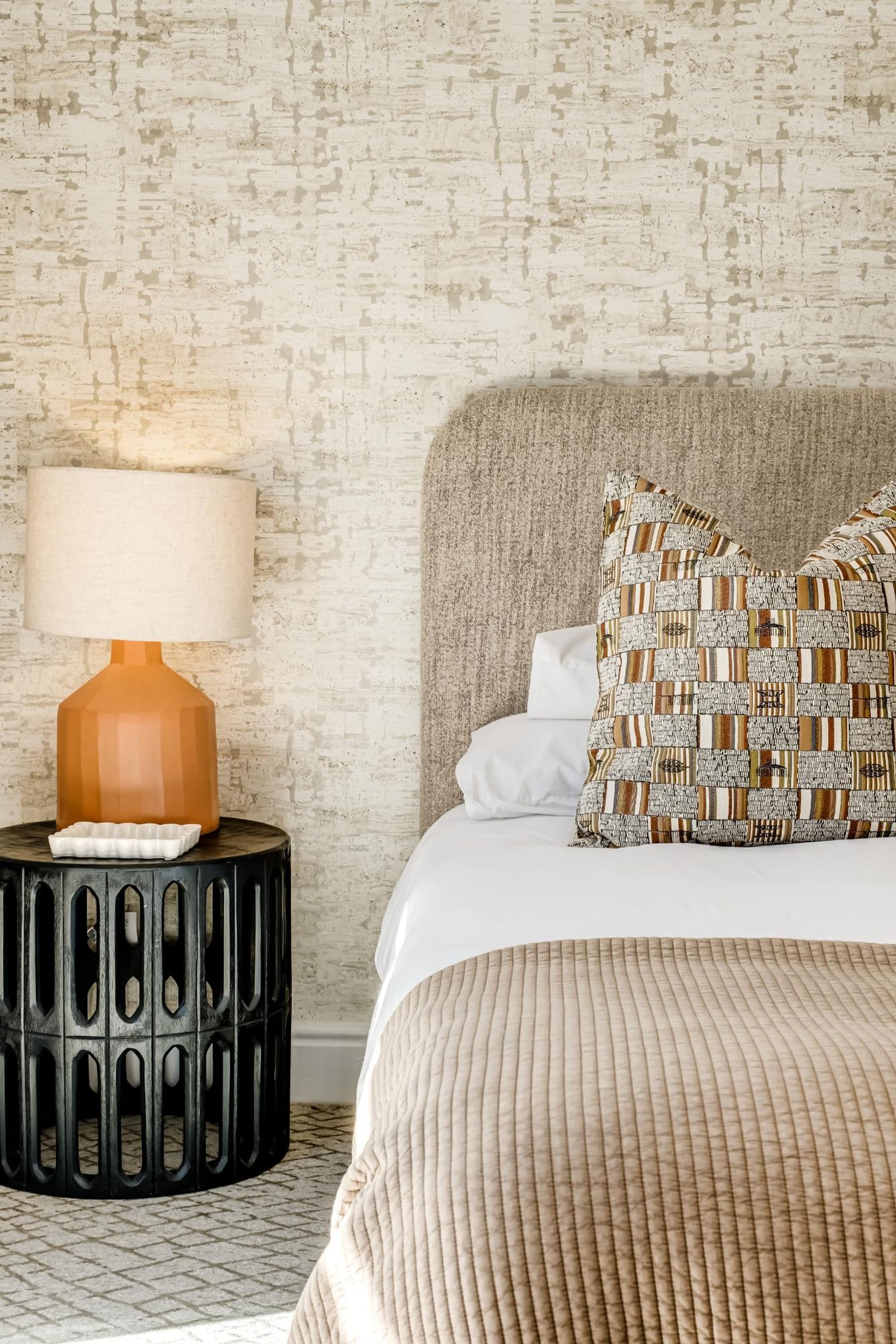 A neatly made bed with patterned pillows, a textured wall, and a lamp on a round side table.