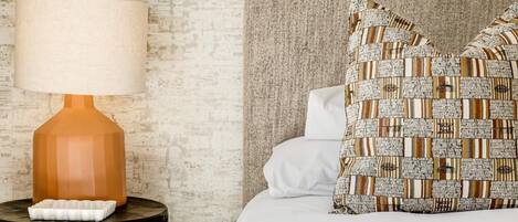 A neatly made bed with patterned pillows, a textured wall, and a lamp on a round side table.