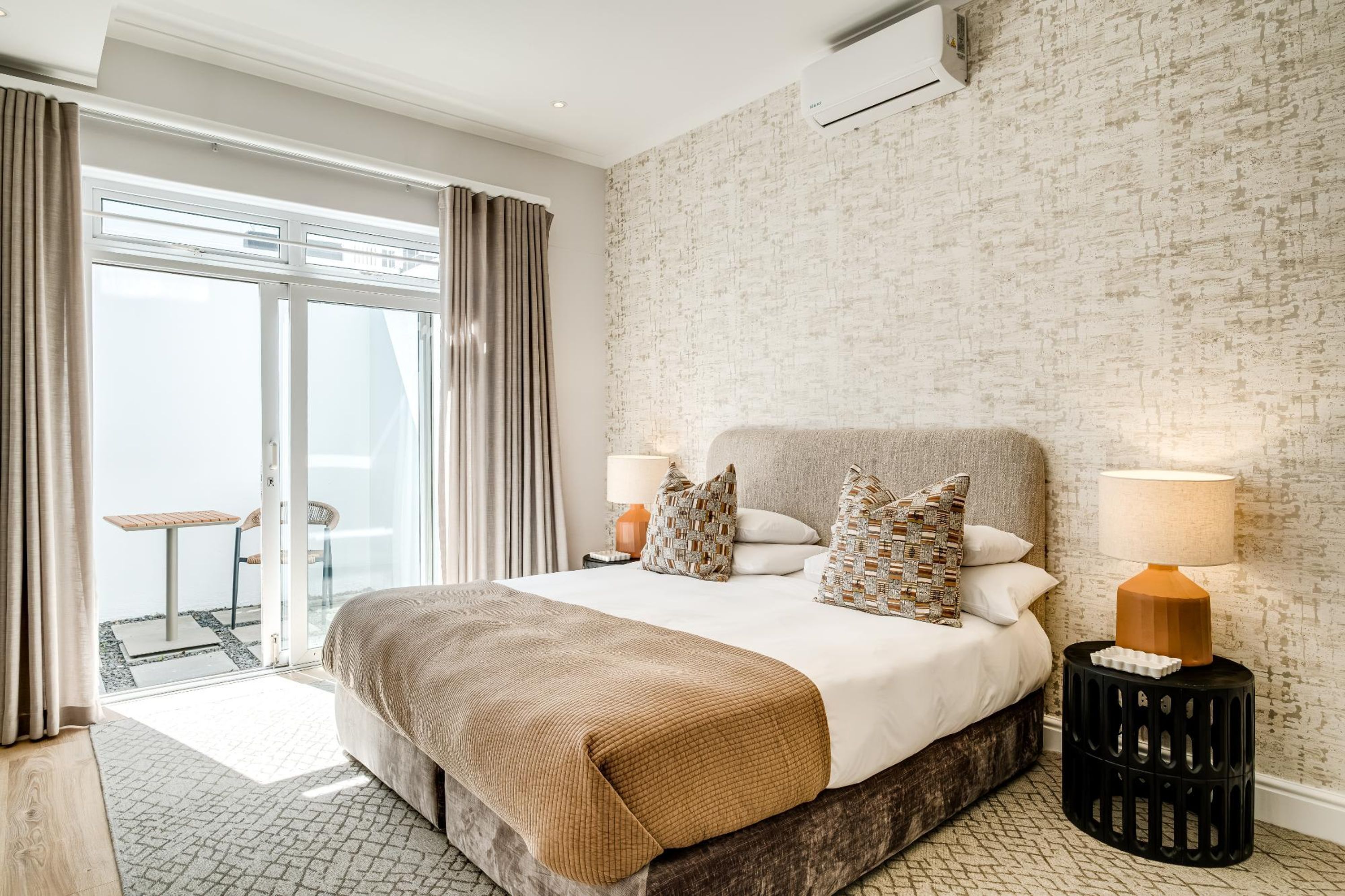 Habitación de hotel moderna con una cama grande, dos lámparas de noche, vista a través de una puerta de cristal de un espacio exterior, y una pared con textura.