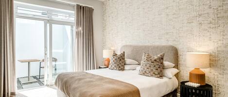 Habitación de hotel moderna con una cama grande, dos lámparas de noche, vista a través de una puerta de cristal de un espacio exterior, y una pared con textura.