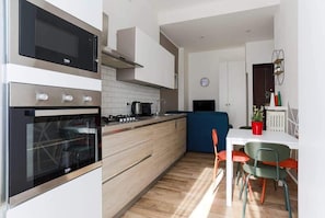 Et moderne køkken med indbygget ovn, mikroovn og komfur. Der er et spiseområde med et hvidt bord og farverige stole.
