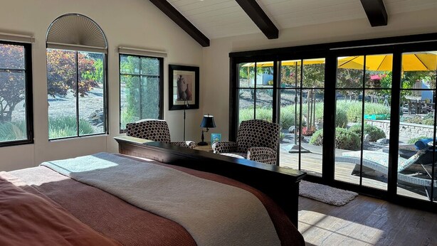 A bedroom with a large bed, a desk, two chairs, and a view of an outdoor pool area through large windows.