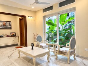 Une pièce avec une table et des chaises blanches, un ventilateur de plafond, une grande fenêtre ornée de plantes et un tableau encadré au mur.