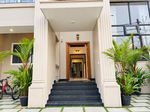 L’entrée d’un hôtel, avec un escalier menant à la porte principale, de part et d’autre duquel se trouvent des plantes en pot.