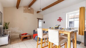 Un comedor con una mesa y sillas de madera, una mesita auxiliar con una planta, y una cocina visible a través de una puerta abierta.