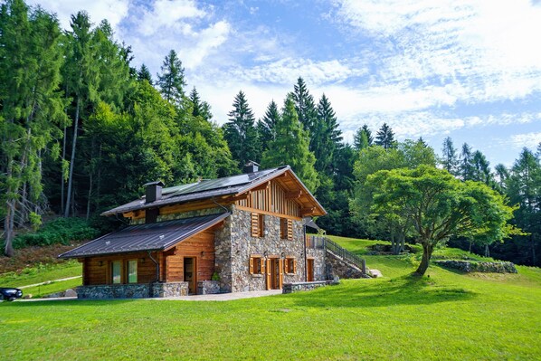 A log cabin with a stone foundation, wooden exterior, and a sloped roof, surrounded by a lush green landscape and tall trees.