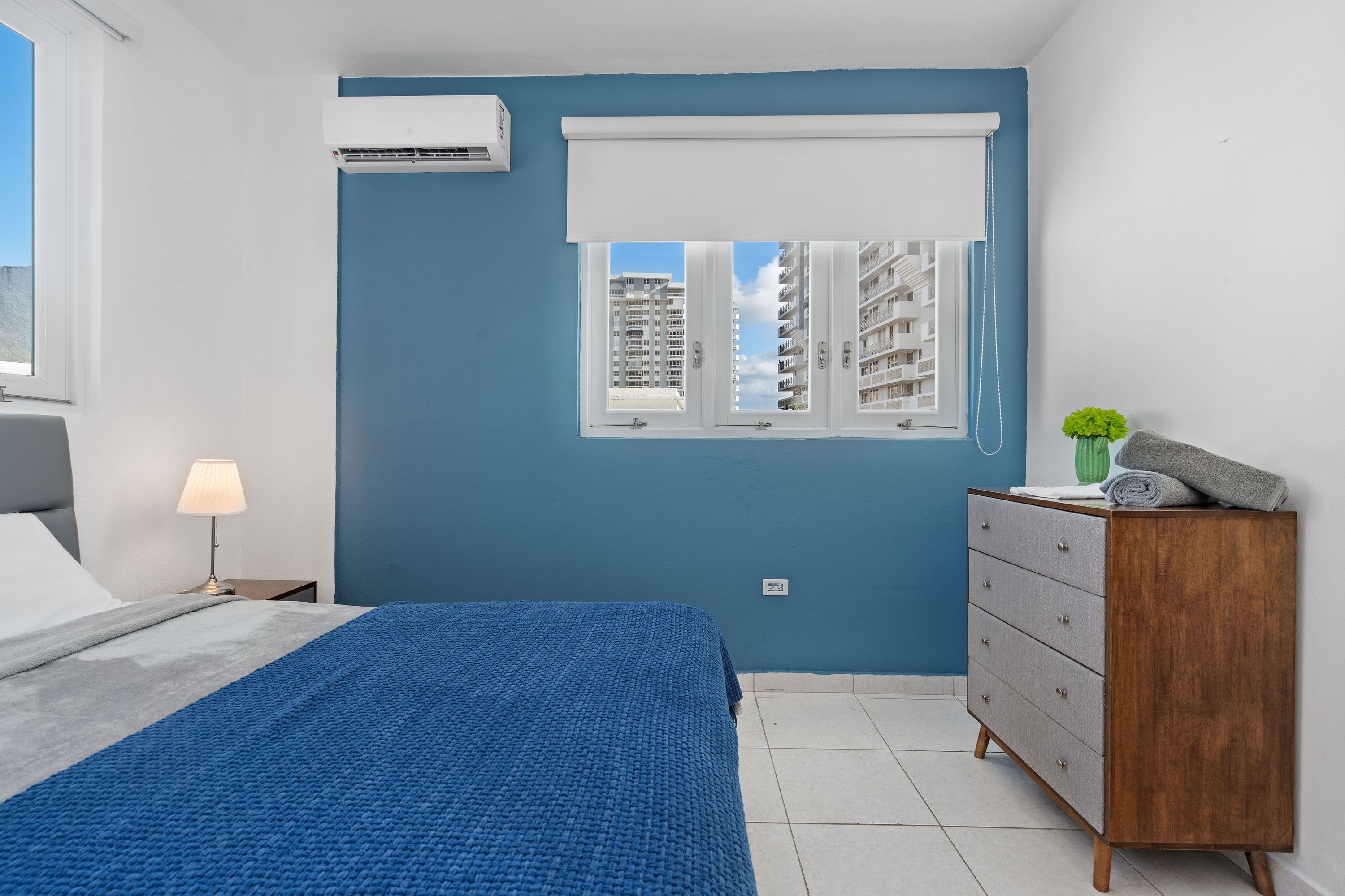 Een slaapkamer met een bed, een dressoir, een raam met jaloezieën en een airconditioning.