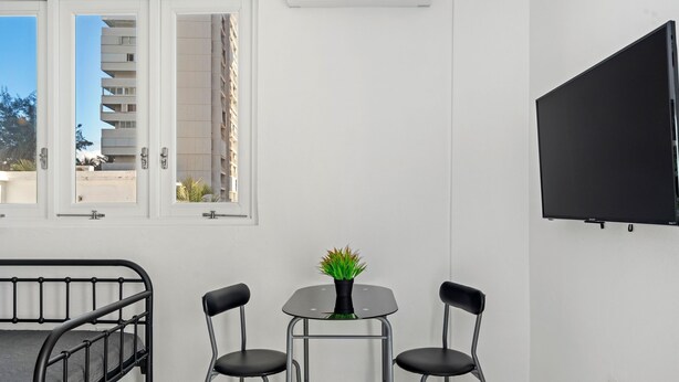 A room with a bed, a small table with two chairs, a potted plant, a TV mounted on the wall, and a window with a view of a building. - Near the Water Shared Pool Pets Permitted (San Juan)