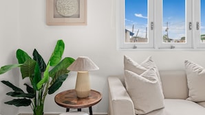 Et værelse med en beige sofa, et rundt træbord med en lampe, en potteplante og et billede i ramme på væggen.
