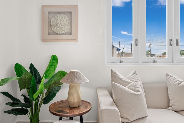 A room with a beige sofa, a round wooden table with a lamp, a potted plant, and a framed artwork on the wall. - Oceanside Swimming Pool Parking (San Juan)