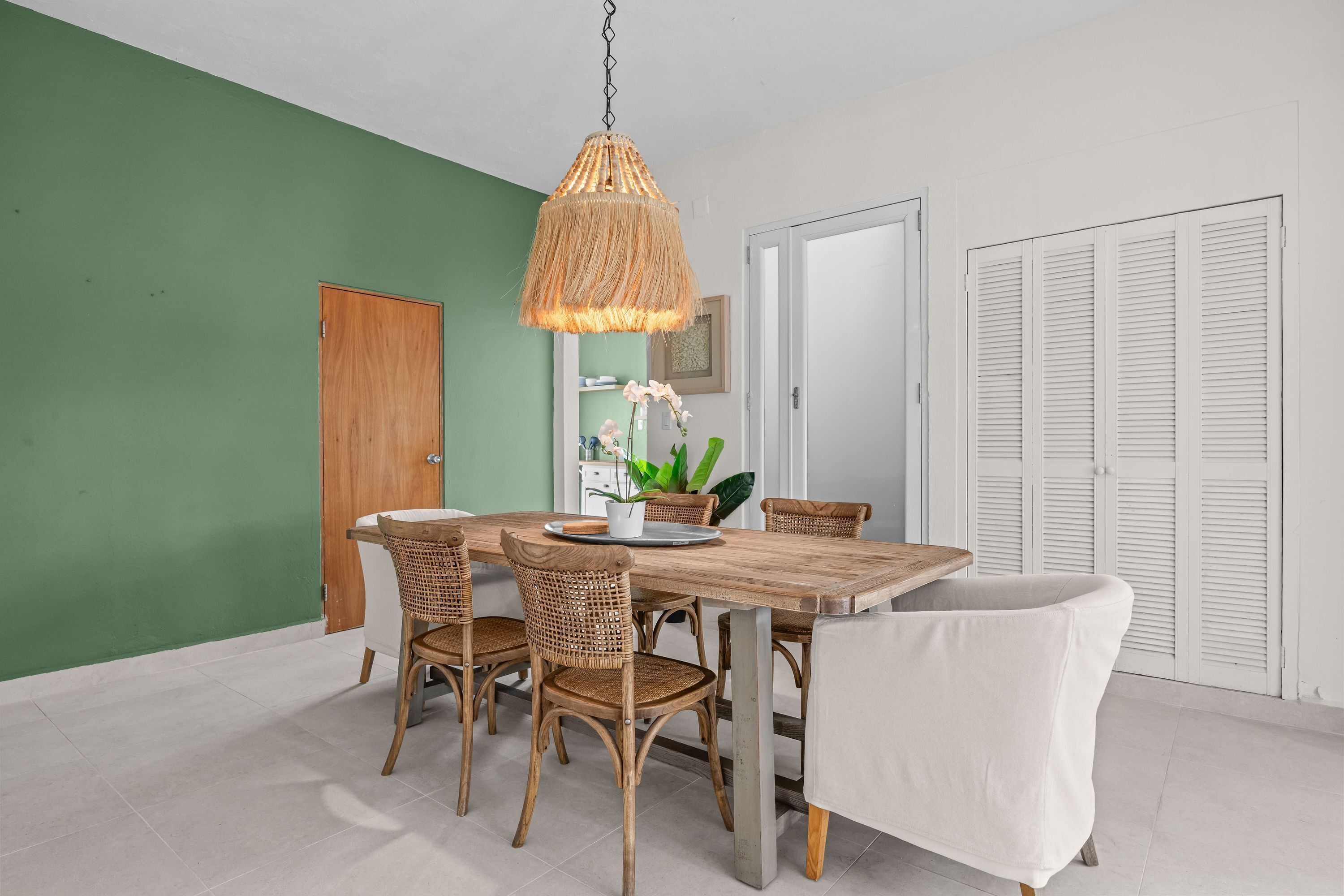 A dining area with a wooden table and wicker chairs, a hanging light fixture, and a white chair.