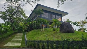 A modern building with a flat roof, surrounded by lush greenery and a large rock. - Palmas - Infinity Pool House - Bagaces La Fortuna. (Provincia de Guanacaste)