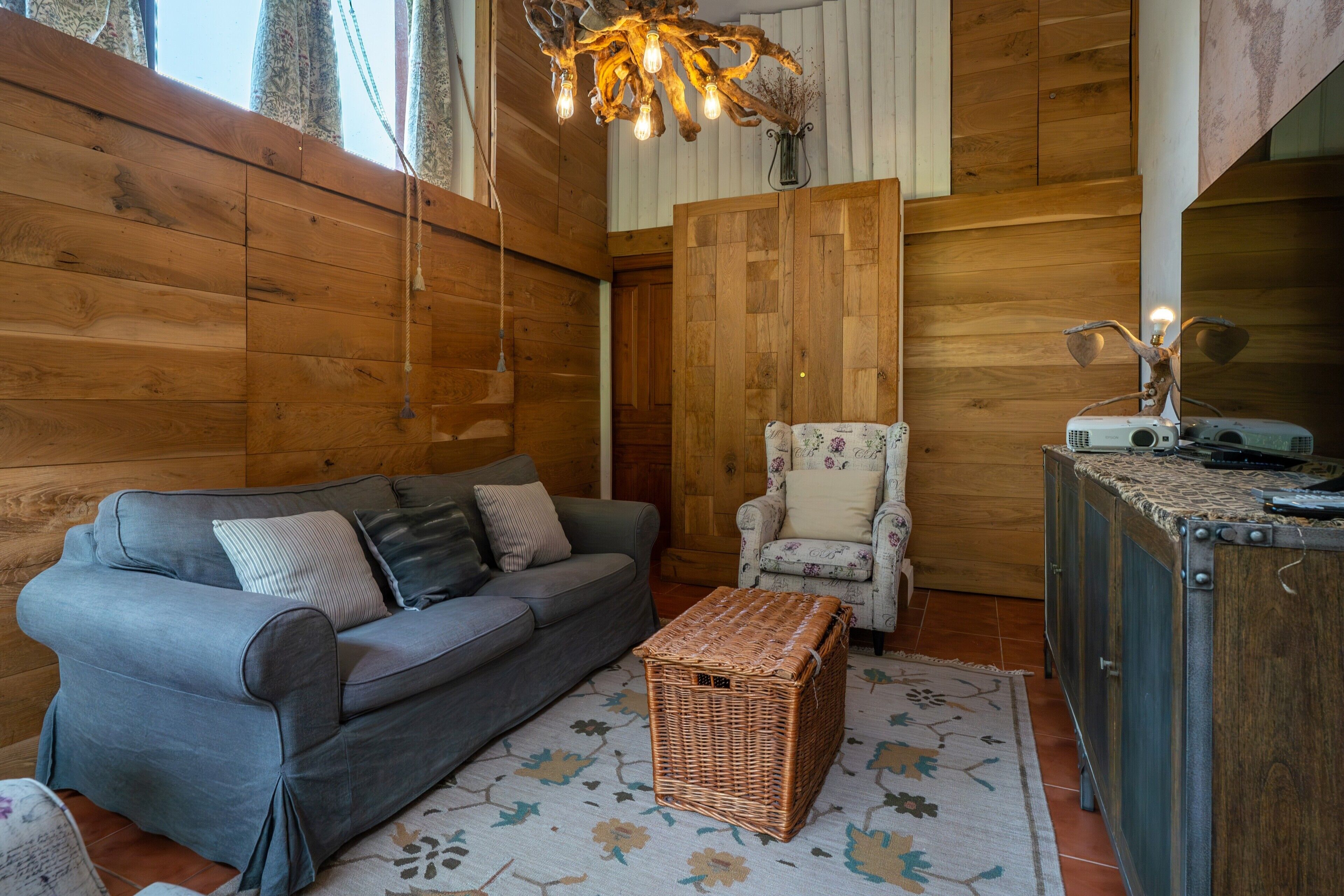 Ein gemütliches Zimmer mit einem grauen Sofa, einem Korbwannenstuhl, einem Blumen-Teppich, einem Holzschrank und einer besonderen Leuchte.