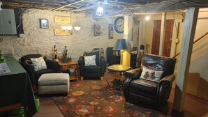 Ein gemütliches Wohnzimmer mit rustikaler Steinwand, einer Uhr und verschiedenen Sitzgelegenheiten, darunter ein Ledersessel und ein kleiner runder Tisch mit einer Lampe.