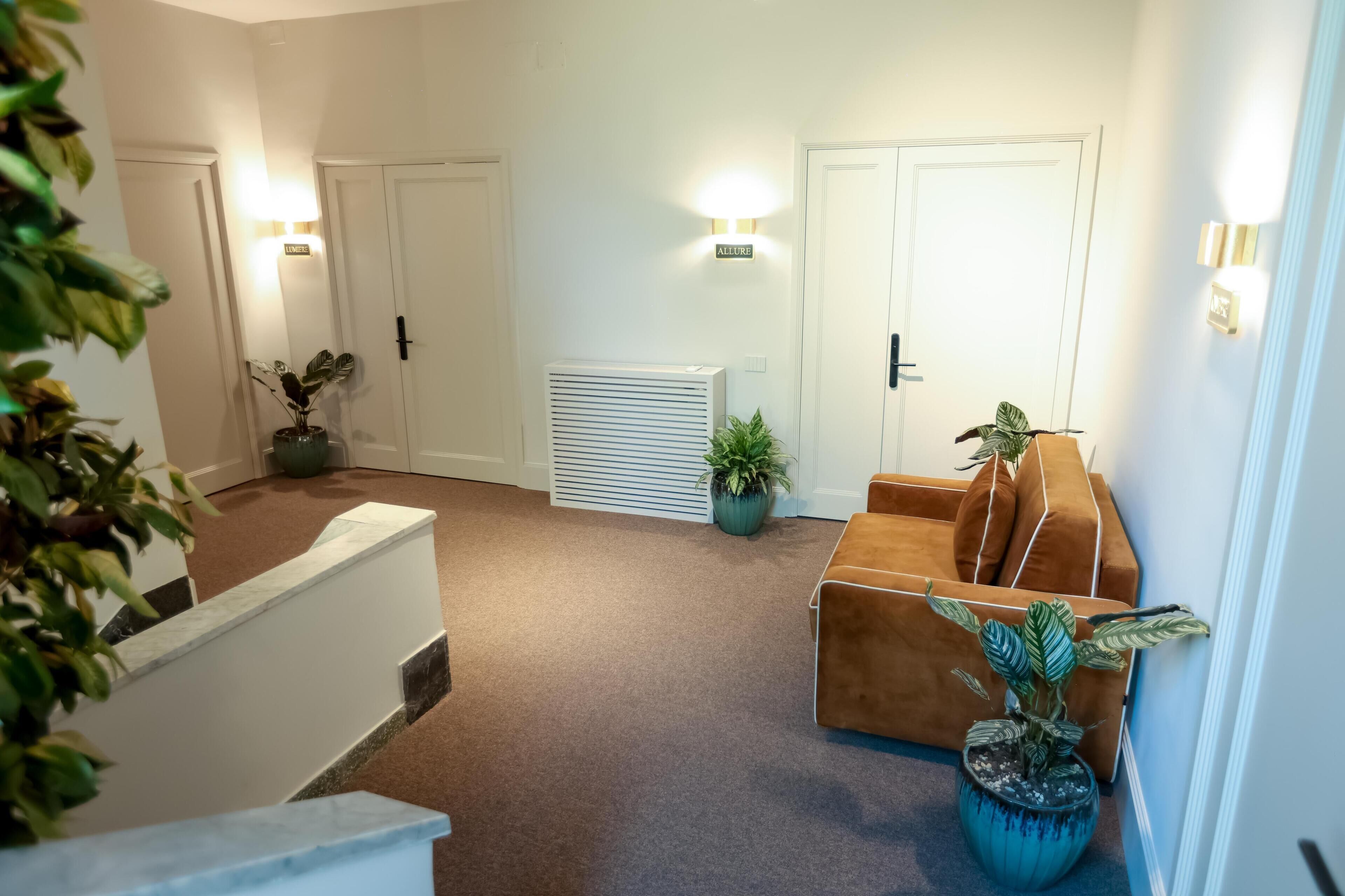 Un vestíbulo de hotel con alfombra marrón, paredes blancas y un sofá marrón. Hay plantas en macetas y un letrero que dice "ALLURE".