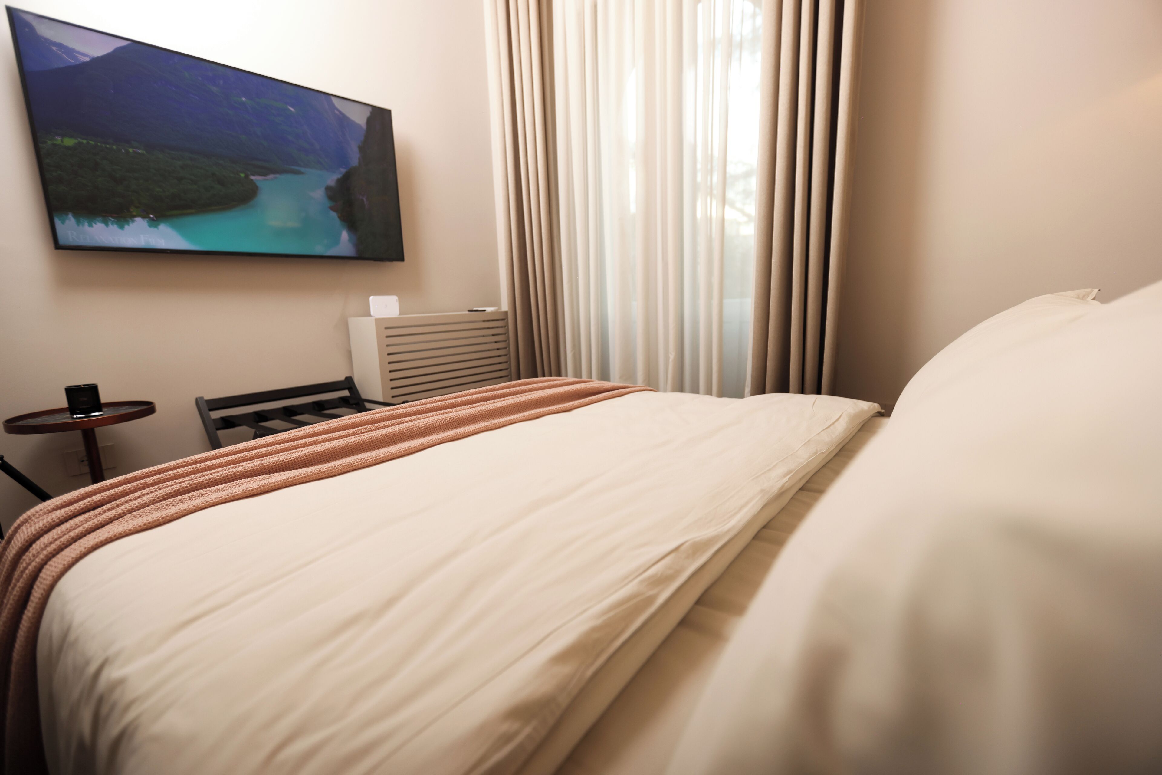 A hotel room with a large bed, a flat-screen TV mounted on the wall, a small table, and a window with curtains.