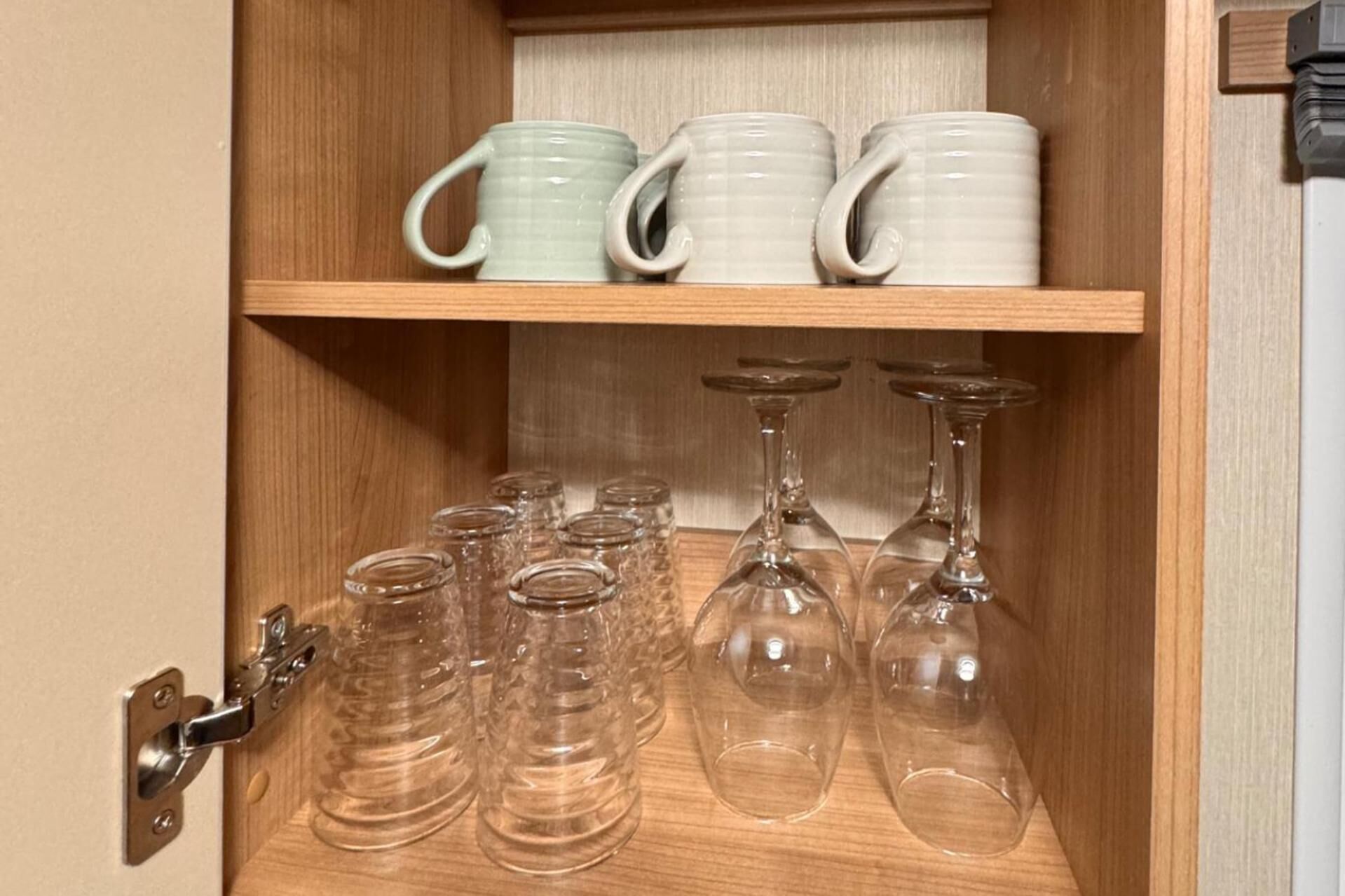 A wooden cabinet with shelves holding clear glassware and ceramic mugs.