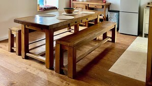 A wooden dining table with benches, a kitchen with a refrigerator, and a window with blinds.