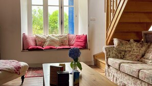 A cozy living room with a wooden ceiling, a window with red checkered curtains, a sofa with floral cushions, and a wooden staircase.