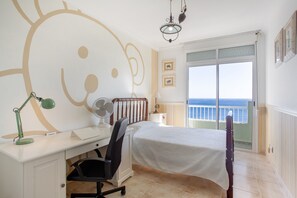 A bedroom with a bed, desk, chair, and a view of the sea through a balcony door.