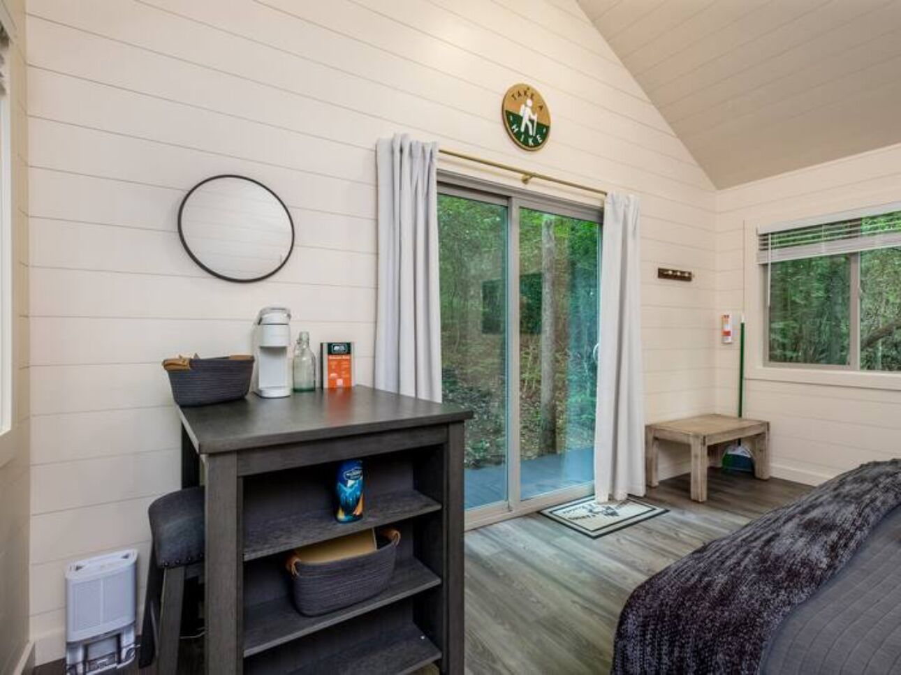A bedroom with a bed, a desk with a coffee maker, a window with curtains, and a view of greenery outside.